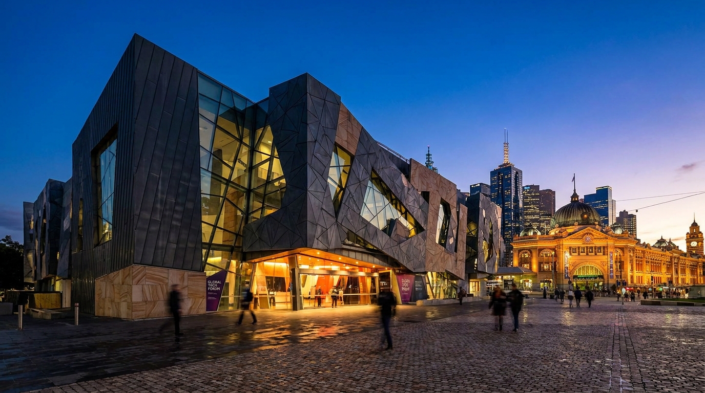 Federation Square in Melbourne at dusk, with the angular glass-and-zinc facade of ACMI lit warmly from within, the Flinders Street Station dome and city skyline behind it.