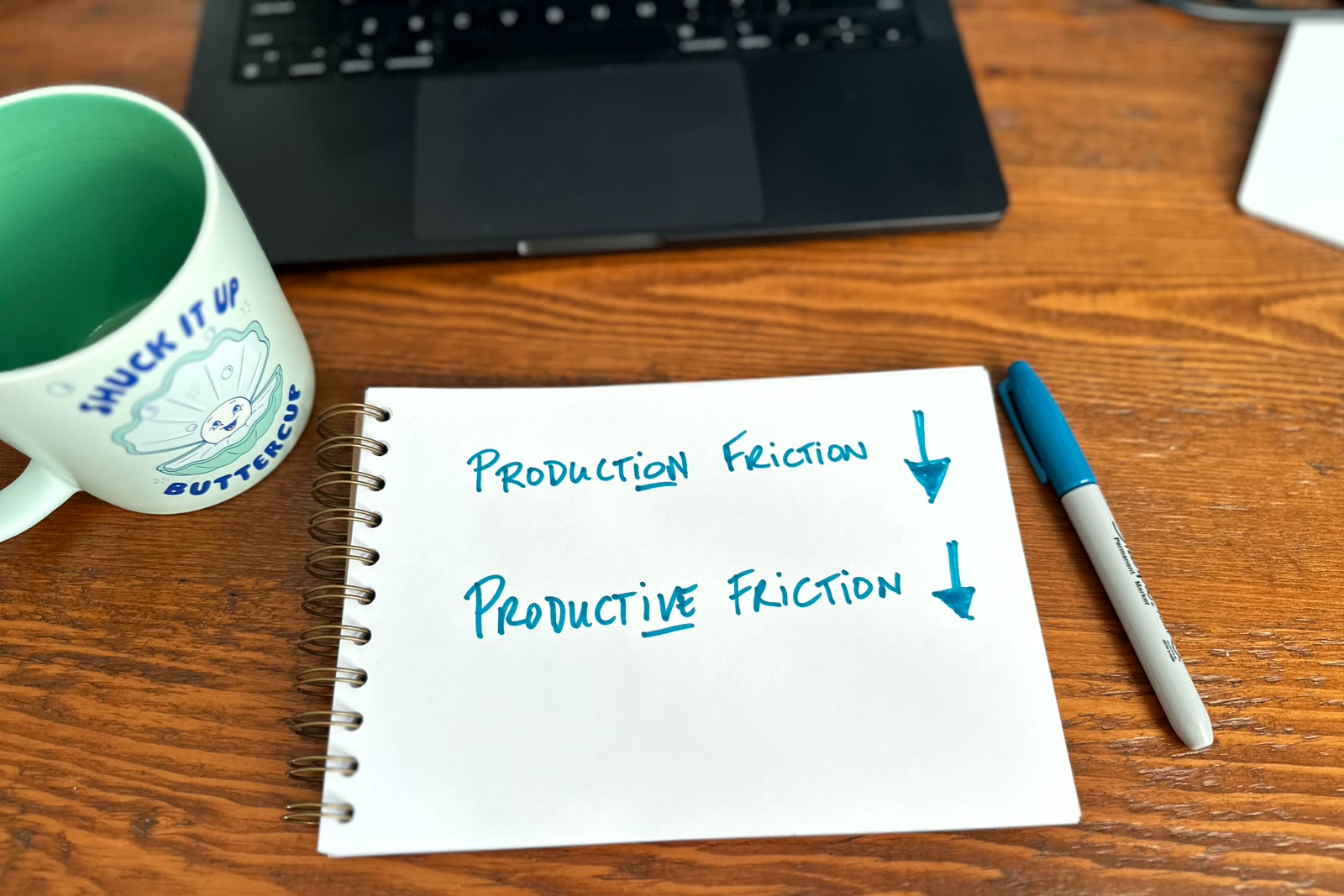 Handwritten in teal marker on a sketchbook page: Production friction, with a large arrow pointing down, and below it Productive friction, with a smaller arrow pointing down. The ion in Production and the ive in Productive are underlined. The sketchbook sits on a wooden desk below an open laptop, between a mint-green mug and the teal marker.
