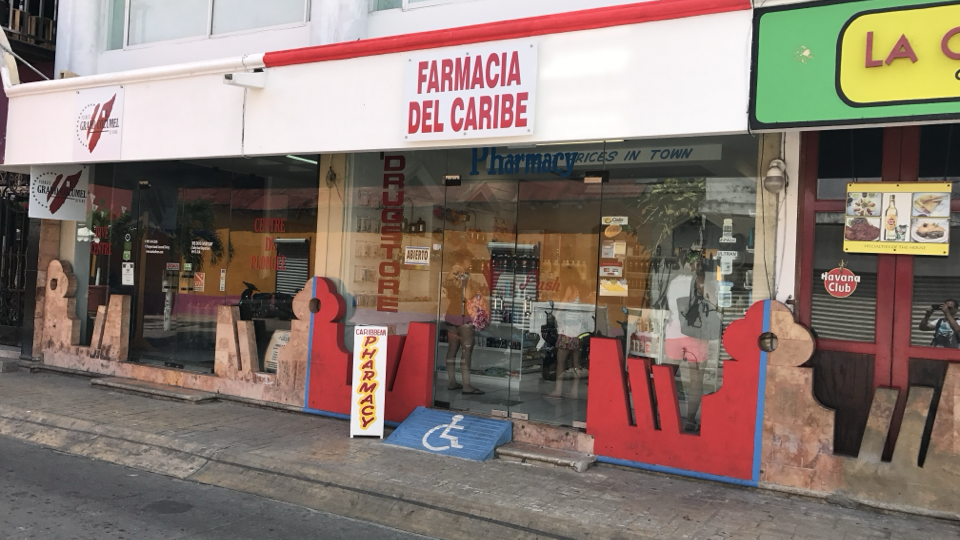 A street photo of the ‘Farmacia del Caribe’ pharmacy in Mexico. A steep makeshift red-and-blue ramp — built for a delivery dolly, not a wheelchair — runs from the doorway straight down to the edge of the road.