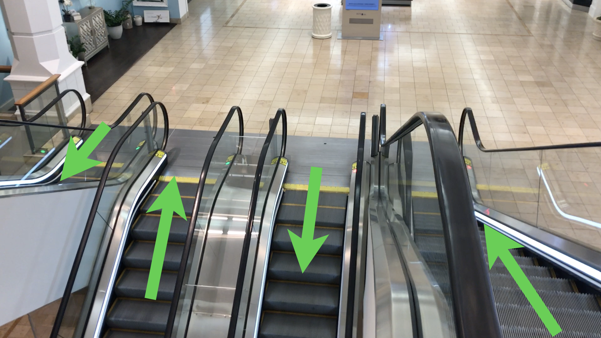 Photo looking down a bank of four side-by-side escalators in a quiet tiled shopping mall. A large green arrow is overlaid on each to show its direction of travel; left to right they read down, up, down, up.
