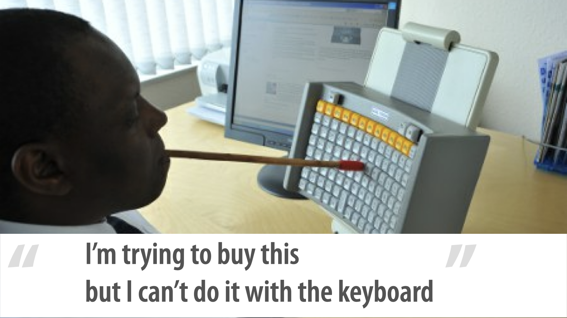 Photo of a man typing with a mouth-wand — a stick held in his mouth — on a compact square grid keyboard angled in front of him, a monitor behind. The grid is tight and curves gently around him so he types by turning his head; the function keys are set off in a different colour. Caption: ‘I’m trying to buy this but I can’t do it with the keyboard.’