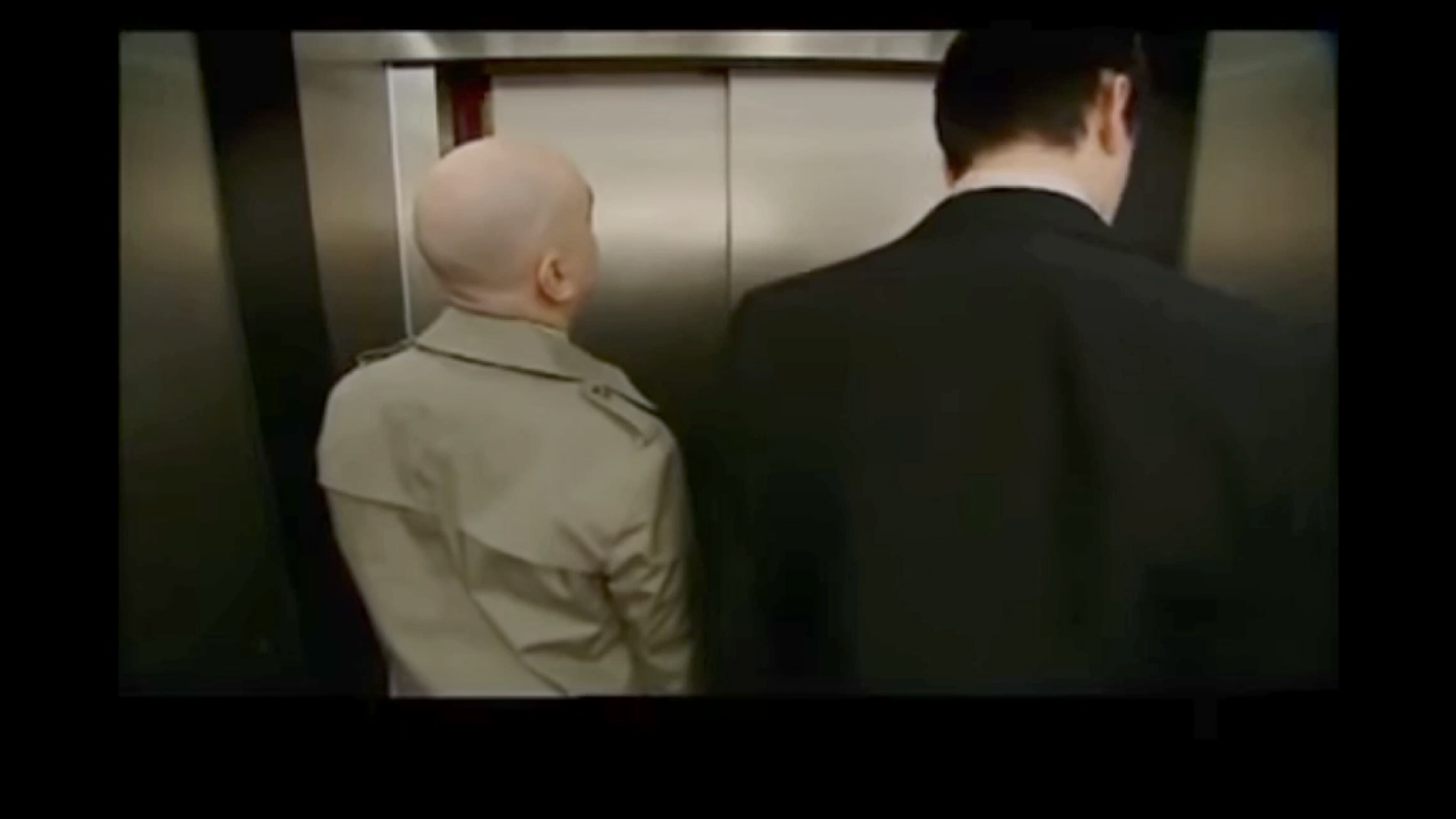 Still from the Burnistoun comedy sketch: two men stand facing the closed doors of a lift — one bald in a tan trench coat, the other in a dark suit.